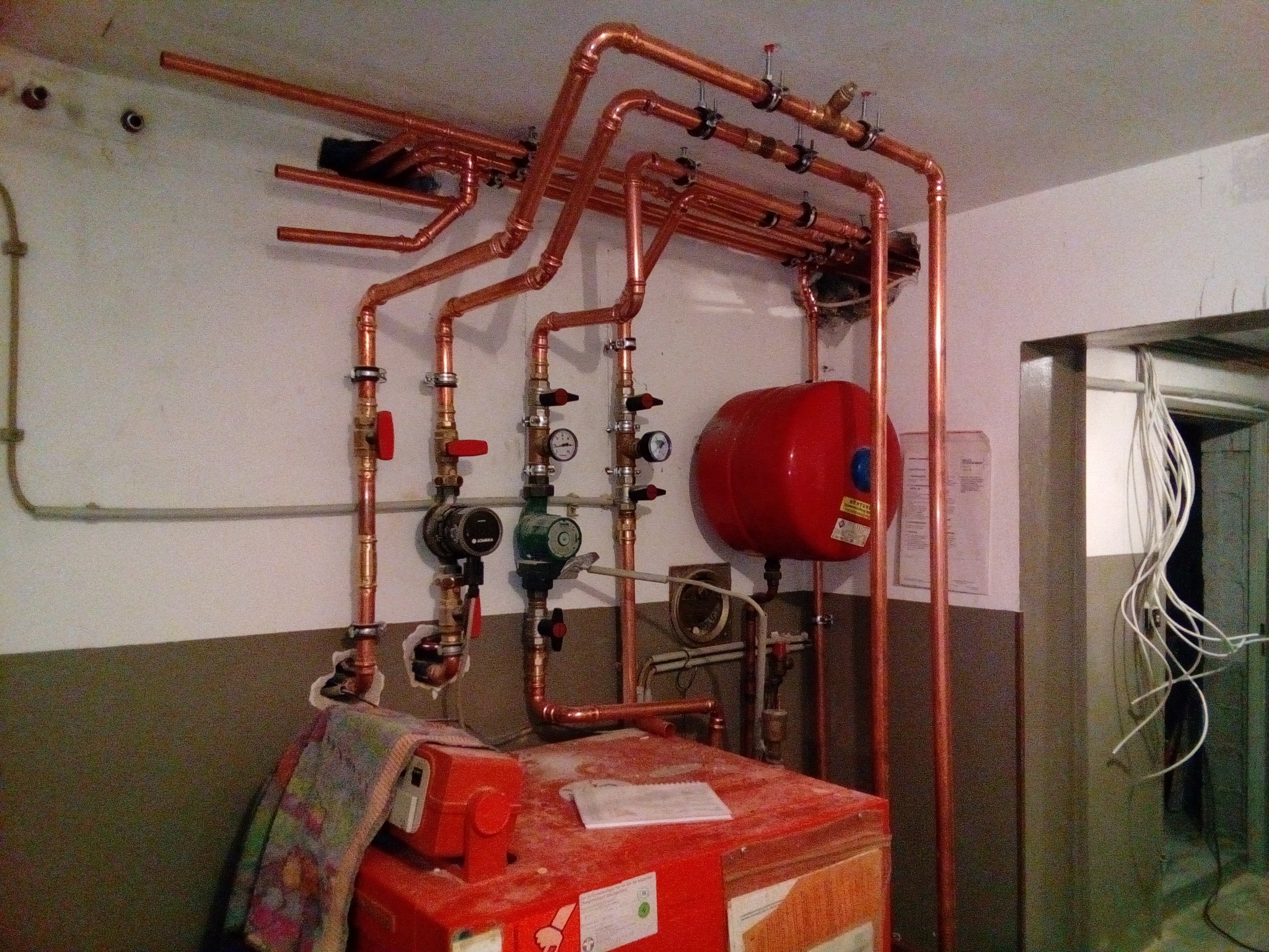 Instalacja centralnego ogrzewania z miedzianymi rurami, manometrami i zaworami, zamontowana na ścianie w pomieszczeniu kotłowni. Na pierwszym planie czerwony piec z ręcznikiem i dokumentami...