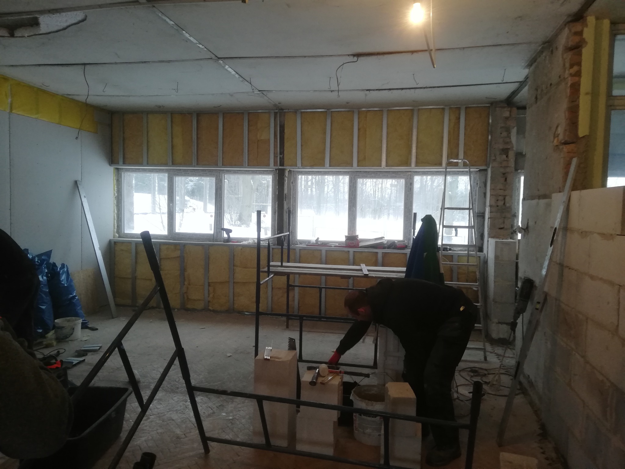 Prace wykończeniowe wewnątrz budynku: montaż izolacji ścian z wełny mineralnej w metalowym stelażu, widoczne narzędzia i materiały budowlane, pracownik w trakcie wykonywania prac murarskich.