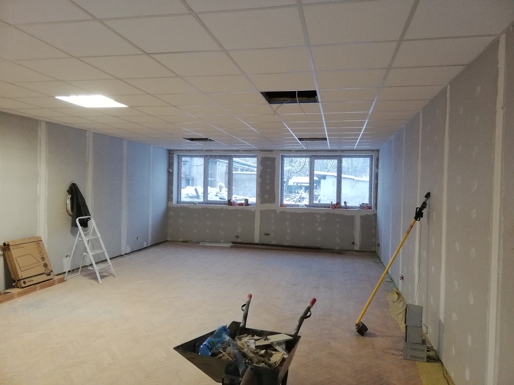 Wnętrze pomieszczenia w trakcie wykańczania: ściany pokryte płytami gipsowo-kartonowymi, sufit podwieszany z otworami, widoczne narzędzia i taczka z odpadami budowlanymi, parkiet na podłodze.