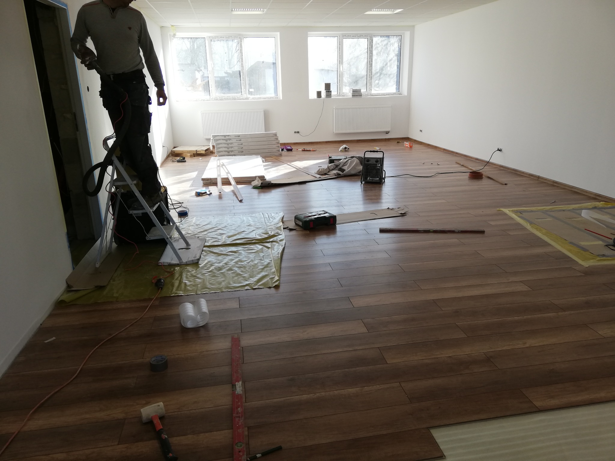 Montaż paneli podłogowych w jasnym, pustym pomieszczeniu; widoczny pracownik na drabinie, narzędzia i materiały przygotowane do układania podłogi.
