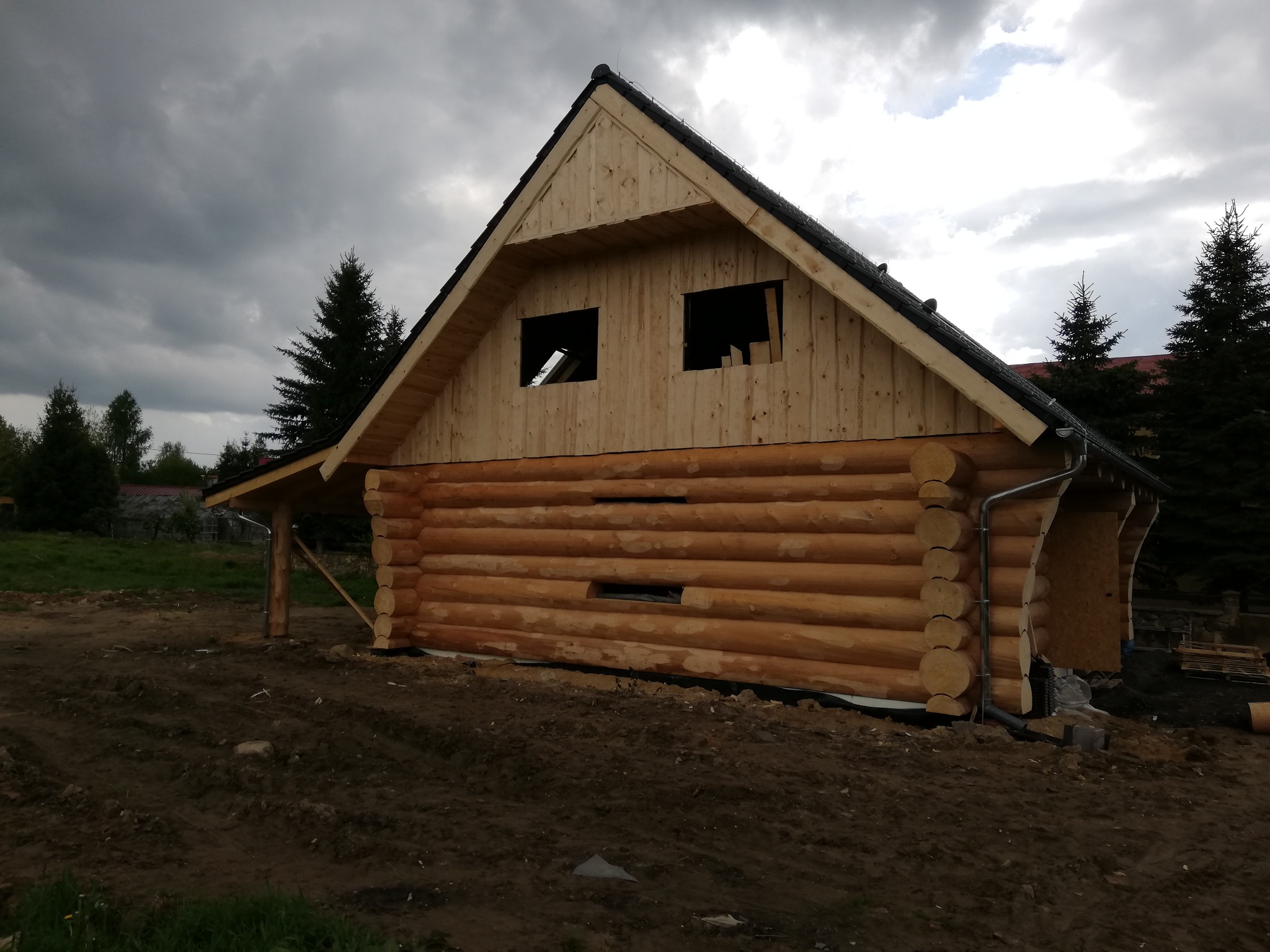 Surowy stan budowy domu z bali drewnianych z widocznym dachem i otworami okiennymi, na tle pochmurnego nieba.