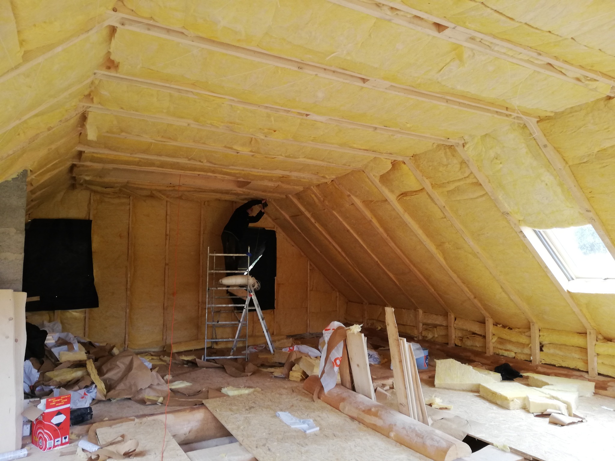 Prace wykończeniowe poddasza: montaż izolacji z wełny mineralnej na drewnianej konstrukcji, widoczny pracownik na drabinie i materiały budowlane na podłodze.