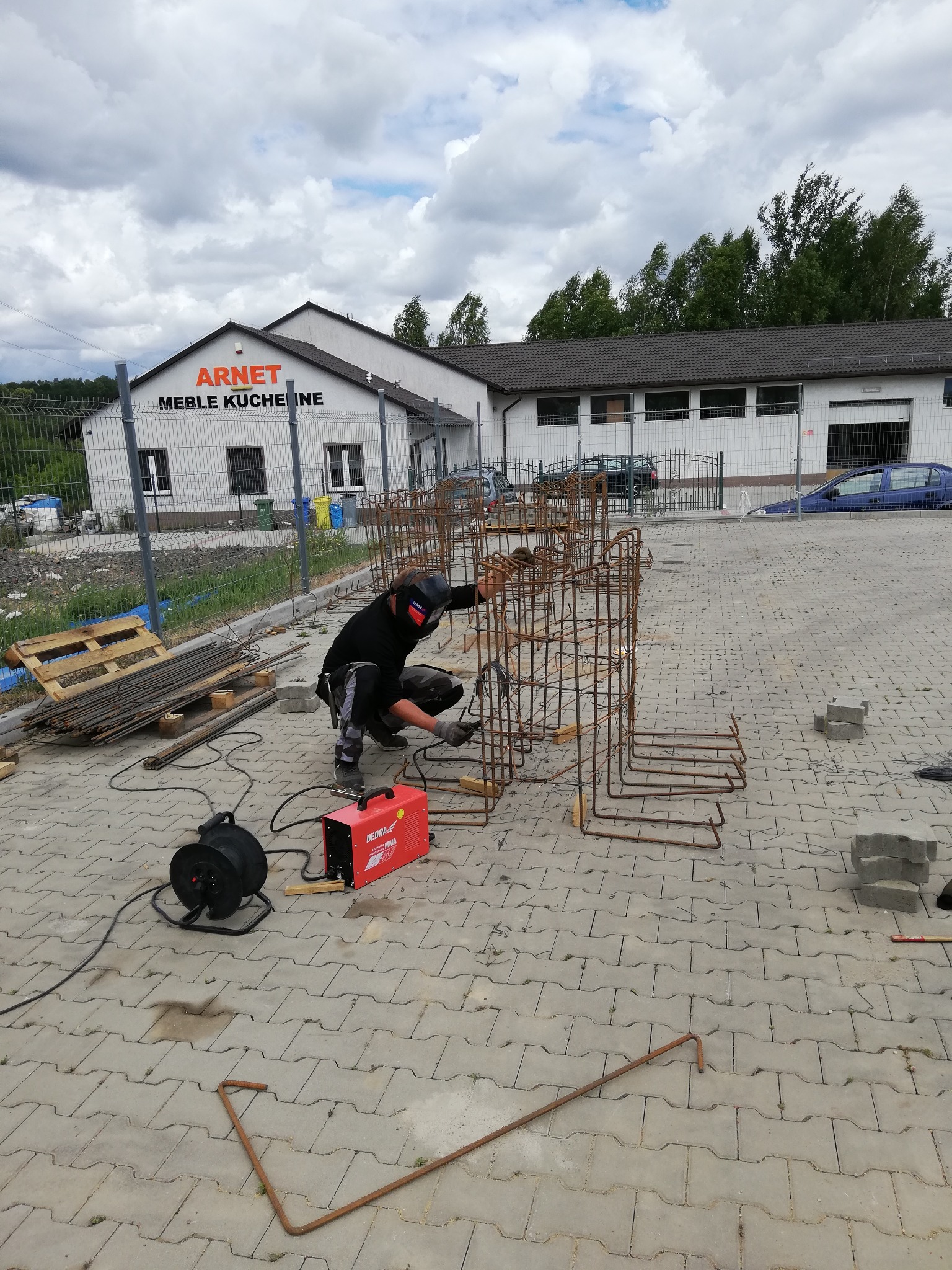 Spawacz w masce spawalniczej łączy zbrojenie fundamentowe na zewnątrz budynku z napisem 'ARNET MEBLE KUCHENNE', używając czerwonej spawarki Dedra MMA.