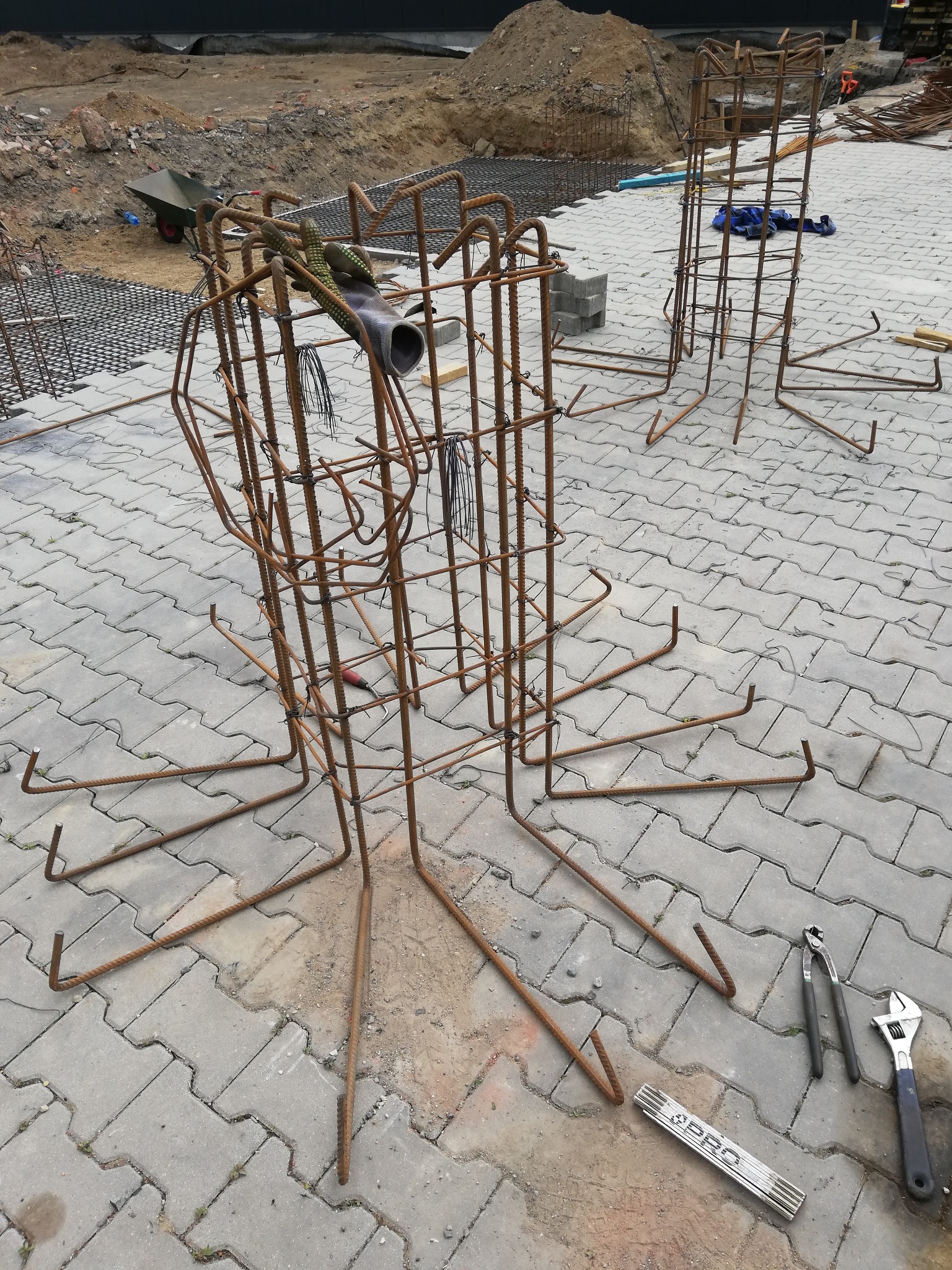 Zbrojenie kolumny fundamentowej z prętów stalowych, przygotowane do zalania betonem na placu budowy, widoczne narzędzia: obcęgi, klucz nastawny i składana miarka.