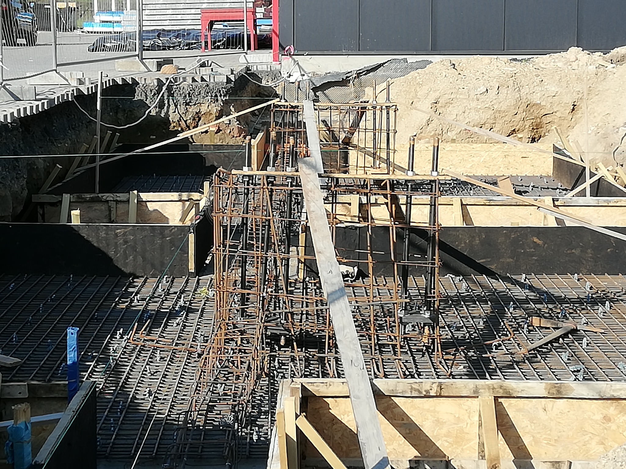 Zbrojenie fundamentów w wykopie, widoczne kosze zbrojeniowe i siatka zbrojeniowa, szalunki z desek i sklejki, ziemia w tle.