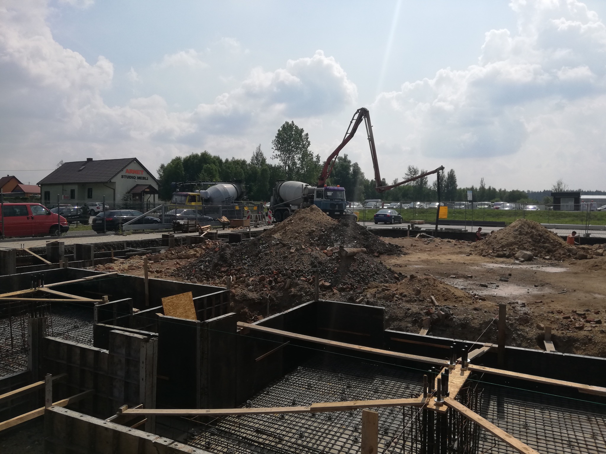 Wykop pod fundamenty z szalunkiem i zbrojeniem, widoczny betonowóz z pompą do betonu oraz hałda ziemi, w tle budynek z napisem Studio Mebli.
