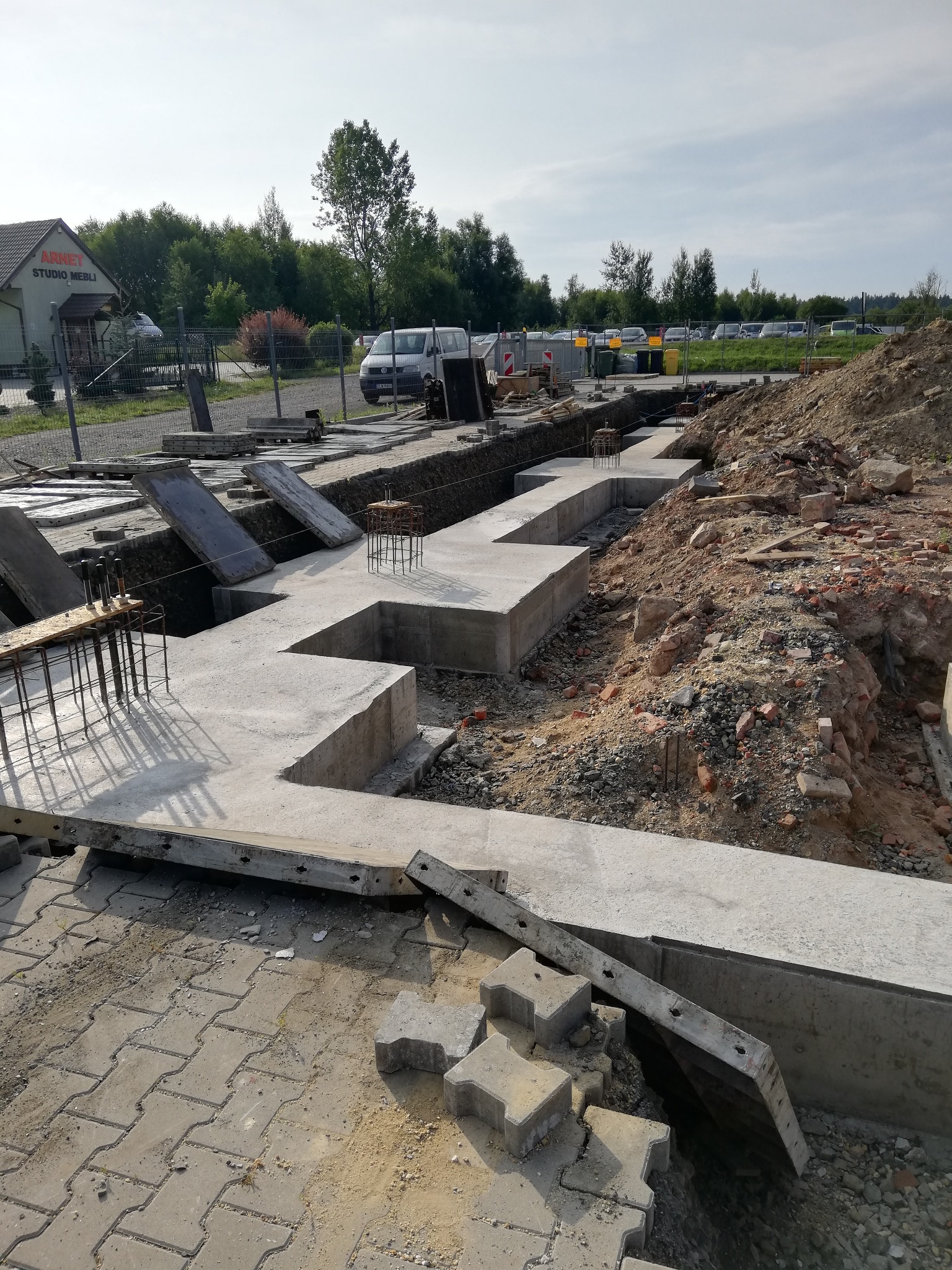 Wylane betonowe fundamenty domu jednorodzinnego z wystającymi prętami zbrojeniowymi, widok z góry na nierówny teren budowy z gruzem i stertą ziemi.