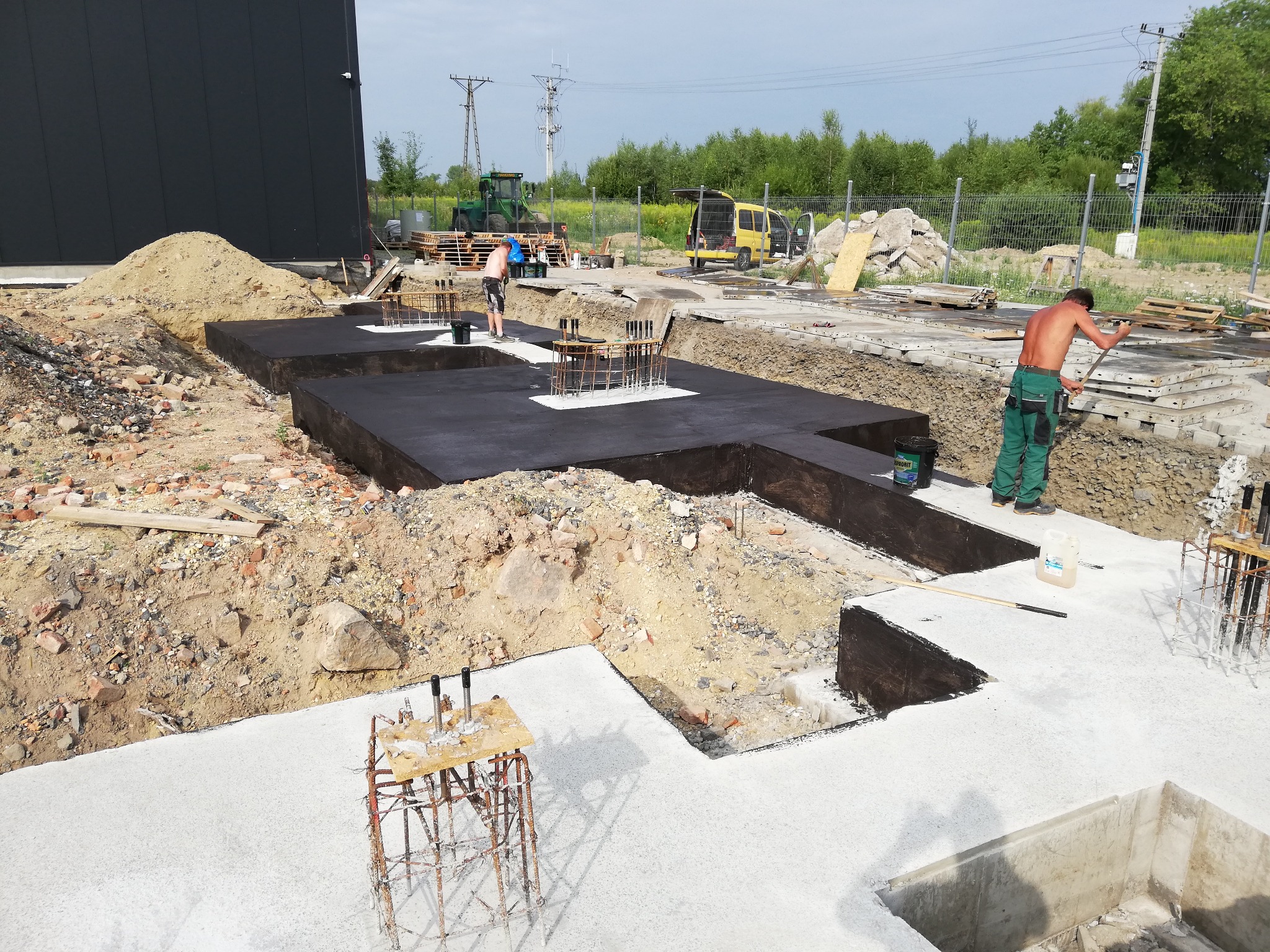 Przygotowanie fundamentów pod budynek: izolacja przeciwwilgociowa na wylanych ławach fundamentowych, zbrojenie wystające, pracownicy na budowie, sprzęt budowlany w tle.