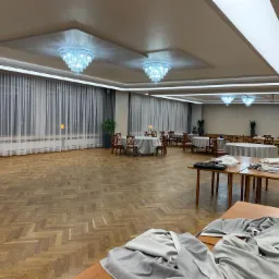 Sala balowa z parkietem w jodełkę, przygotowywana do wydarzenia: stoły z obrusami, krzesła i kryształowe żyrandole podwieszane pod sufitem. Na pierwszym planie stoły z materiałem dekoracyjnym.