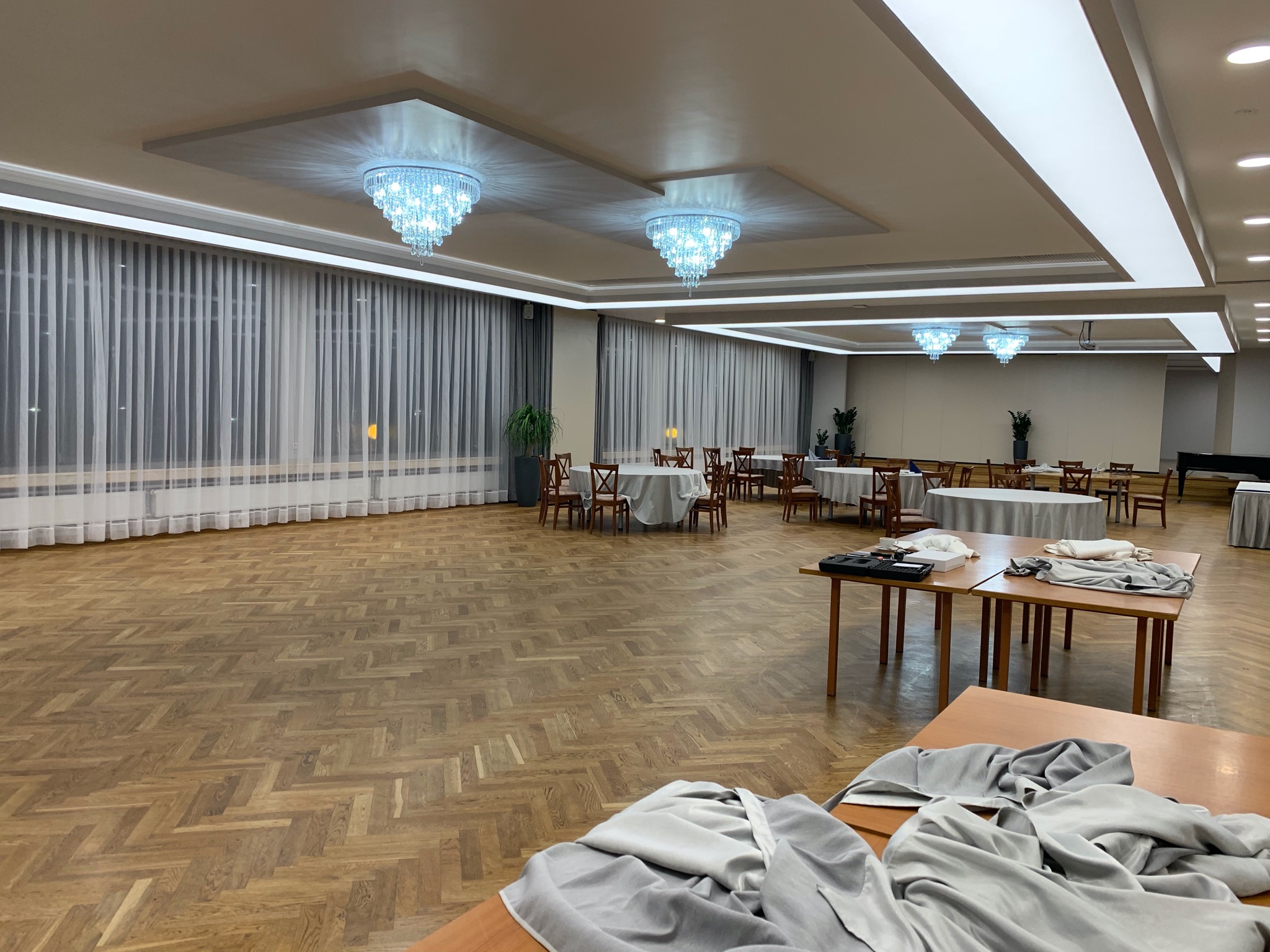 Sala balowa z parkietem w jodełkę, przygotowywana do wydarzenia: stoły z obrusami, krzesła i kryształowe żyrandole podwieszane pod sufitem. Na pierwszym planie stoły z materiałem dekoracyjnym.