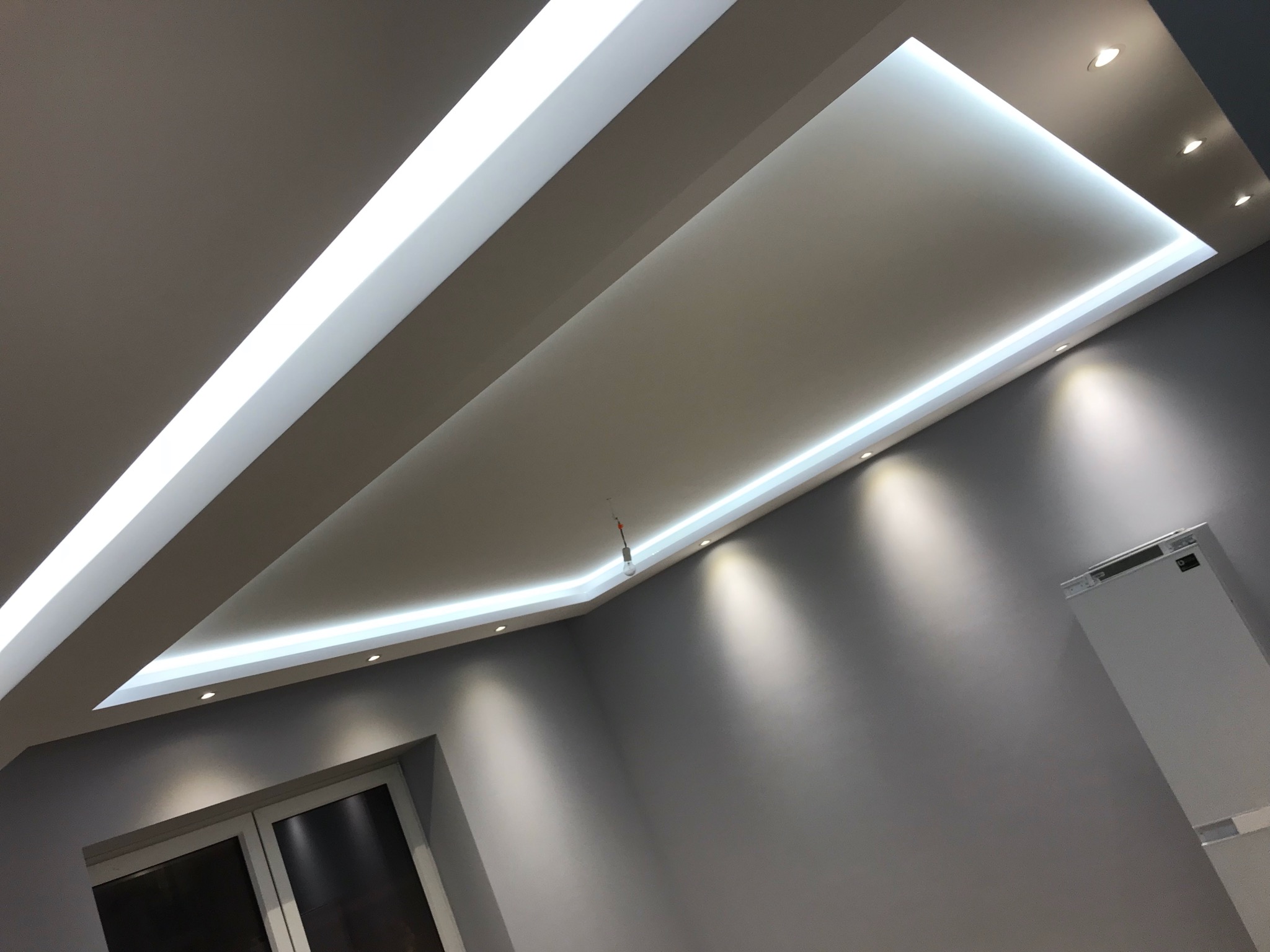 Nowoczesny sufit podwieszany z oświetleniem LED w kształcie prostokąta, punktowe lampy halogenowe, szare ściany i okno w dolnym rogu kadru.