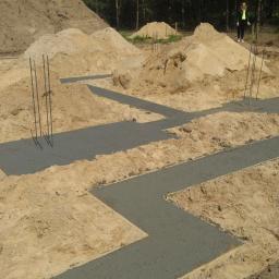 KnoppersBud - Wylane fundamenty pod dom szkieletowy z wystającymi prętami zbrojeniowymi, otoczone piaskiem i zalesionym terenem w tle, widoczna osoba w oddali.