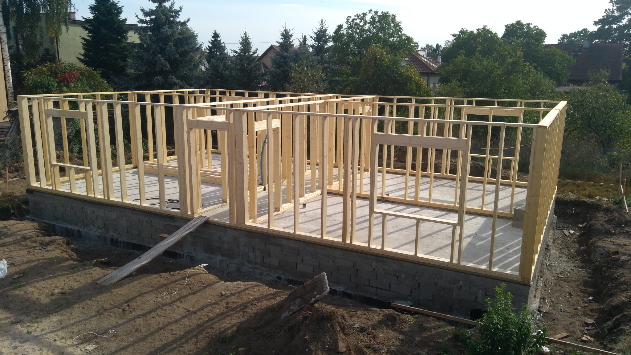 Wznoszenie konstrukcji budynku z drewnianych belek na fundamencie z bloczków betonowych, widoczne otwory okienne i drzwiowe, w tle zieleń drzew.