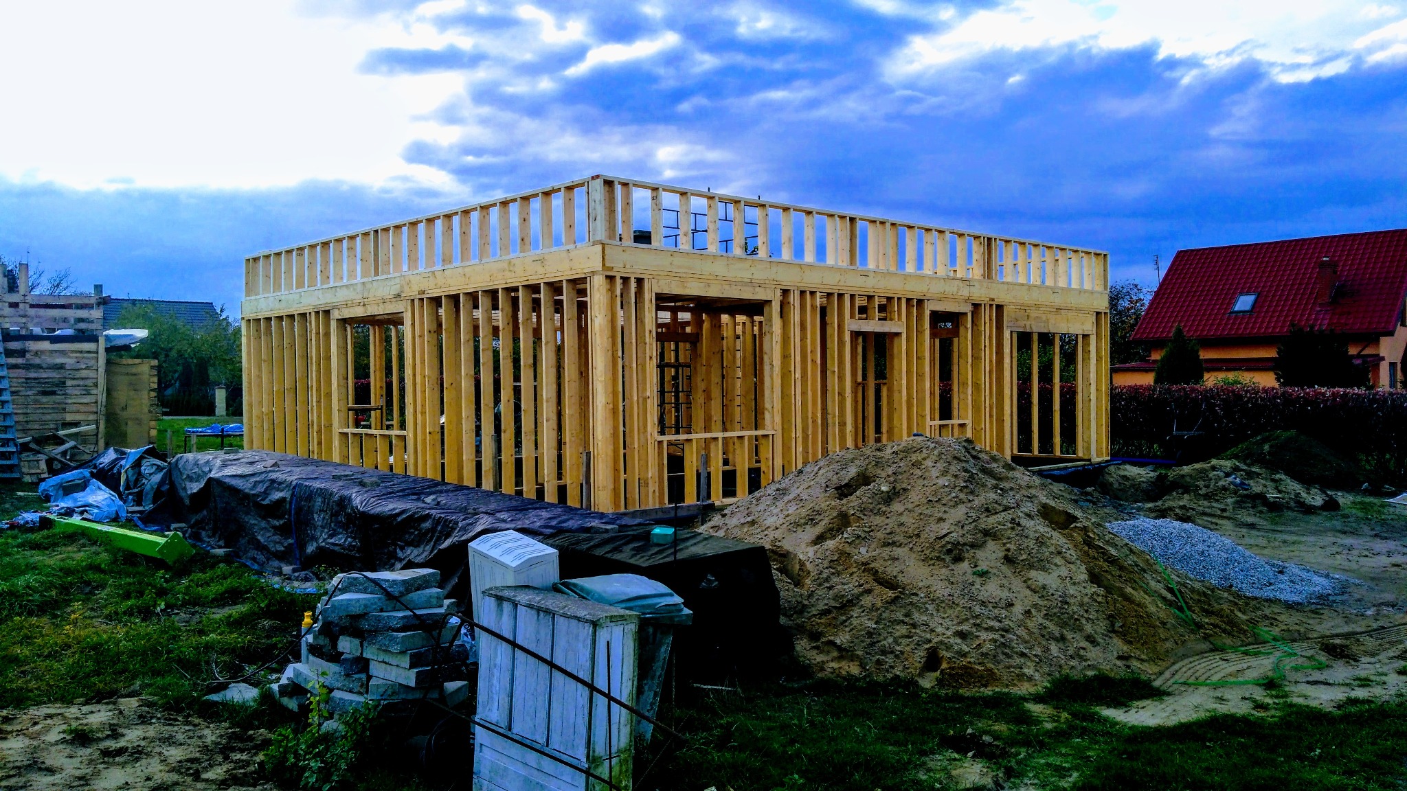 Wznoszenie konstrukcji drewnianej domu w technologii szkieletowej, widoczny szkielet ścian i stropu, na pierwszym planie hałda piasku i gruzu, w tle dom z czerwoną dachówką.