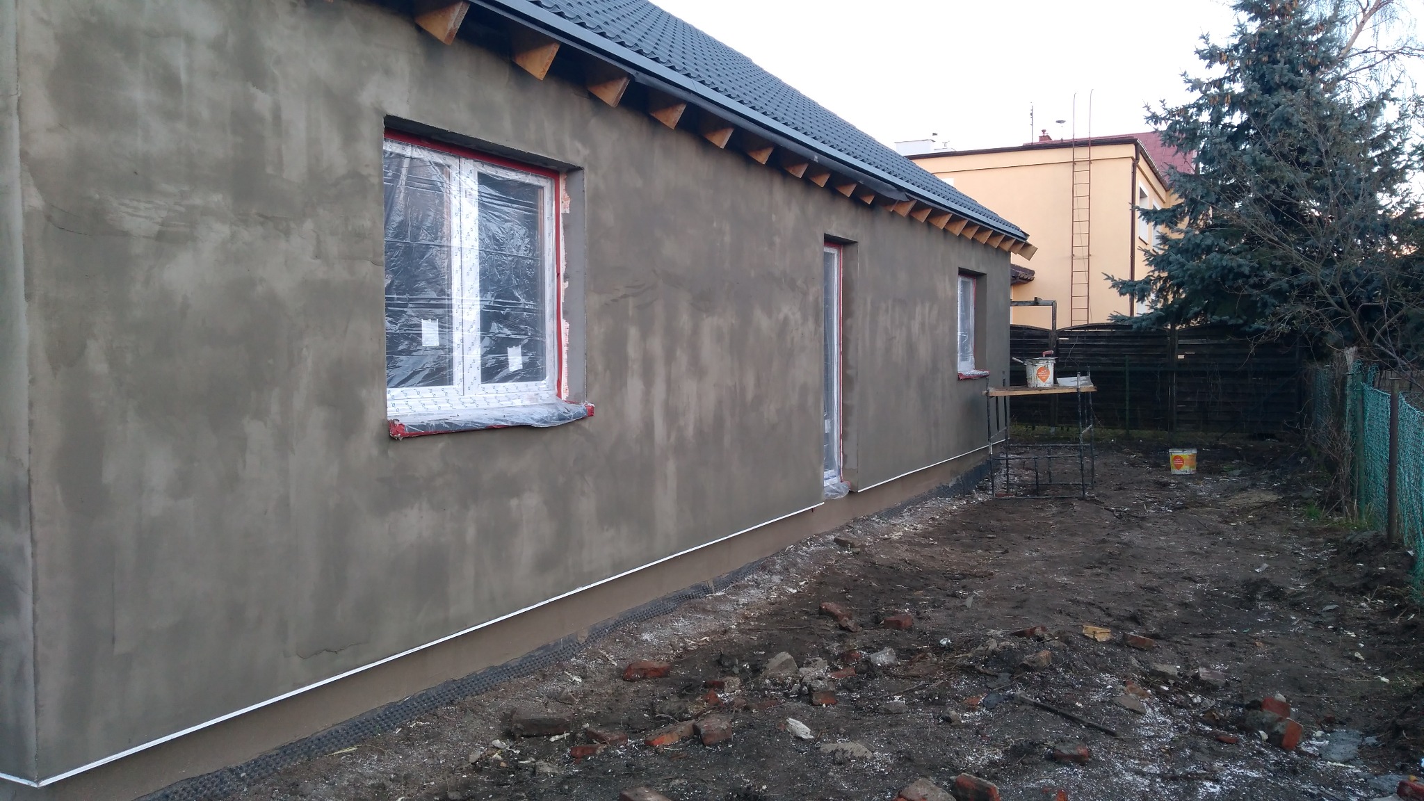 Elewacja domu w trakcie prac wykończeniowych, okna zabezpieczone folią, widoczne rusztowanie z farbami, teren wokół domu częściowo rozkopany.