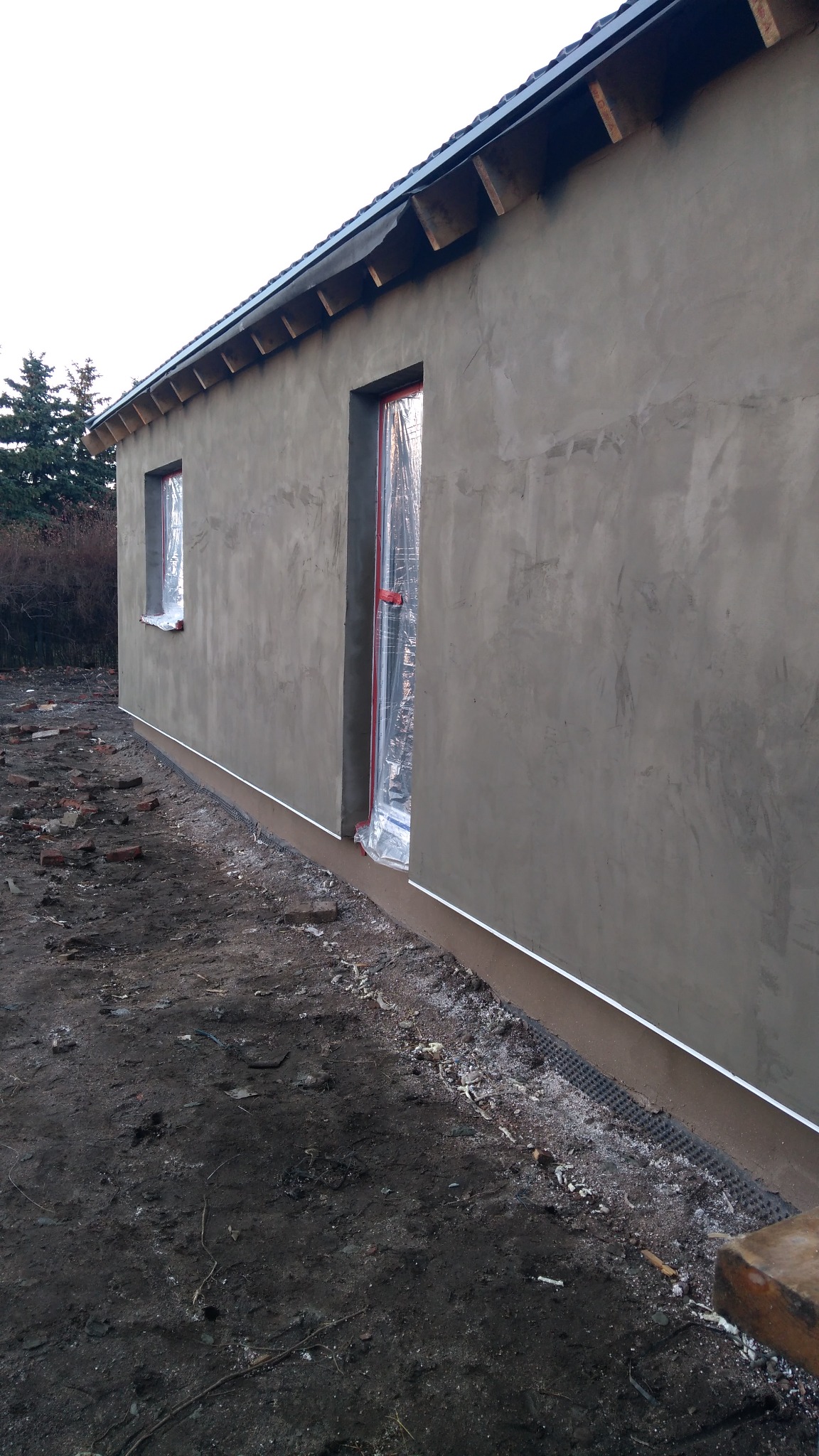 Surowa elewacja budynku w trakcie budowy, okna i drzwi zabezpieczone folią, widoczna podmurówka i przygotowany teren wokół.