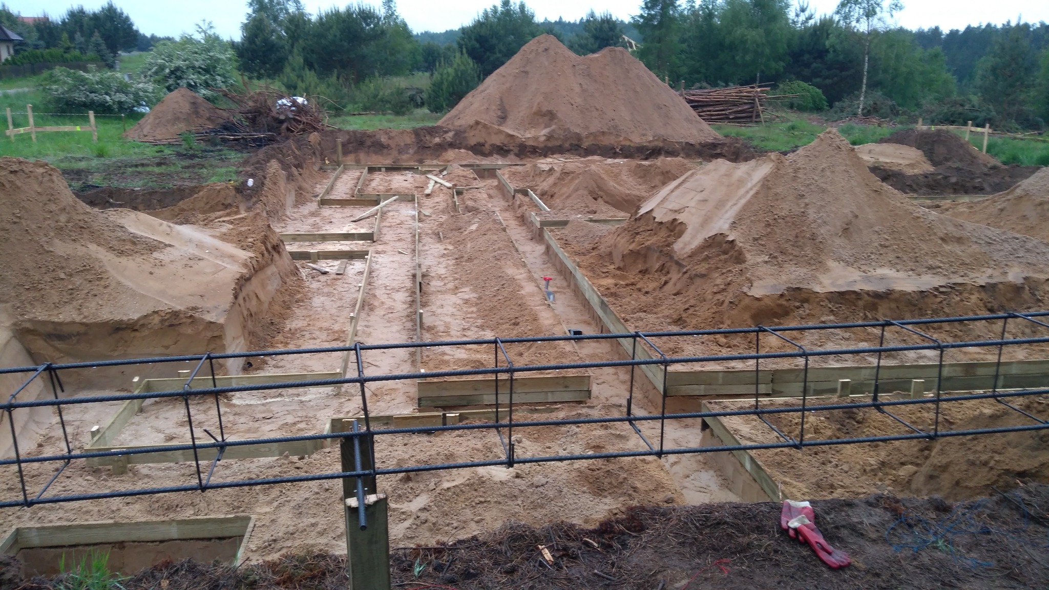 Wykonane wykopy pod fundamenty domu z widocznym szalunkiem i zbrojeniem, w tle usypane pryzmy ziemi.