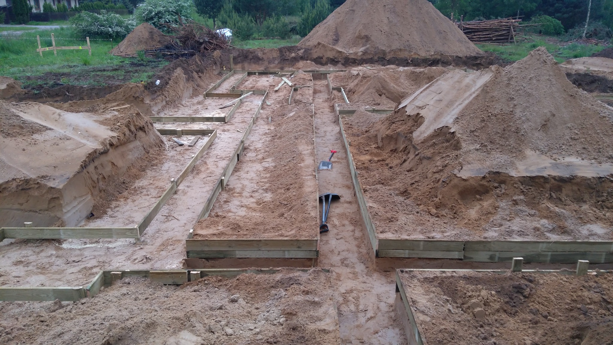 Wykop fundamentów z drewnianym szalunkiem, hałdy ziemi w tle, łopata i kilof leżące w wykopie.