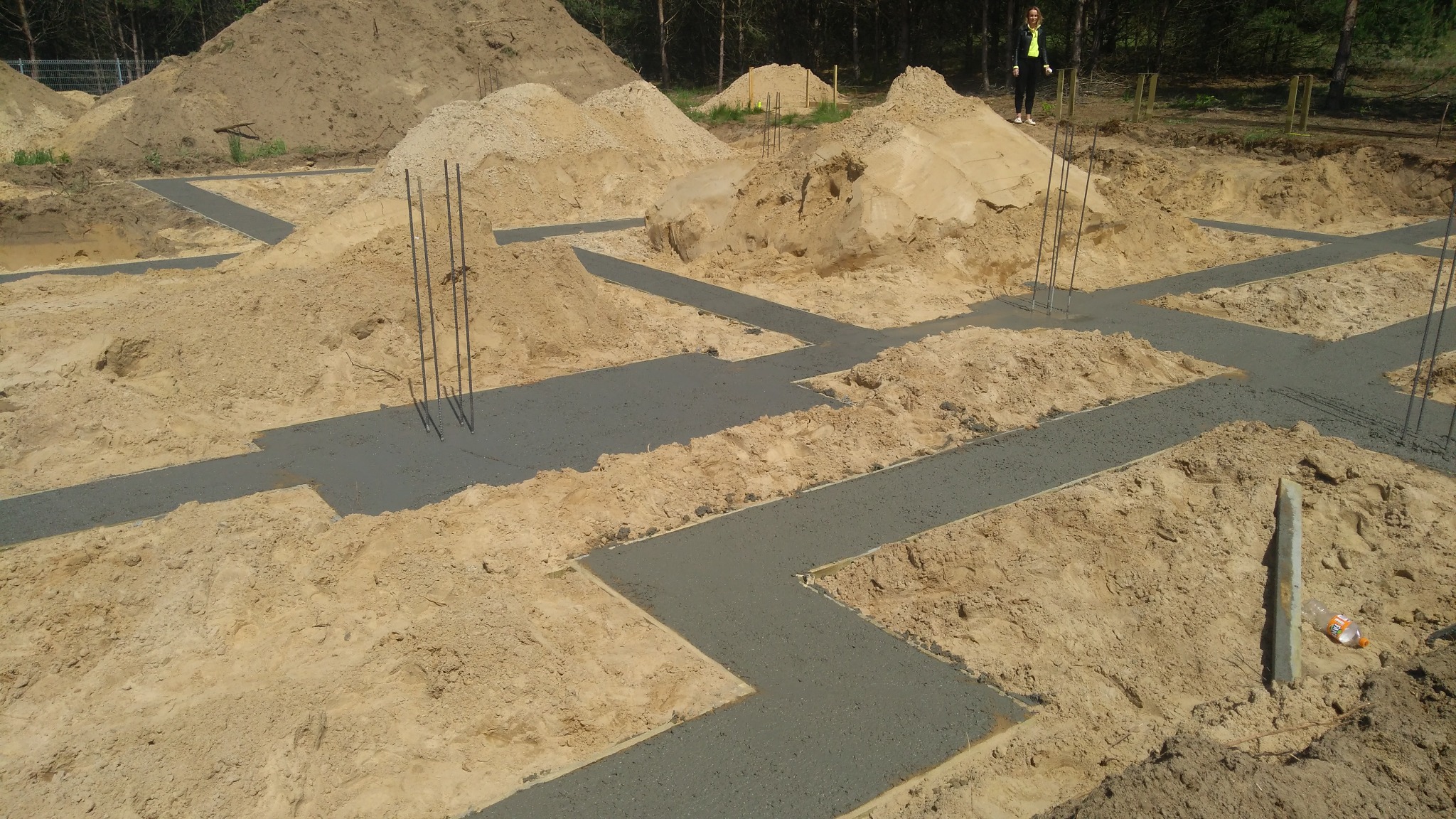Wylane fundamenty pod dom szkieletowy z wystającymi prętami zbrojeniowymi, otoczone piaskiem i zalesionym terenem w tle, widoczna osoba w oddali.