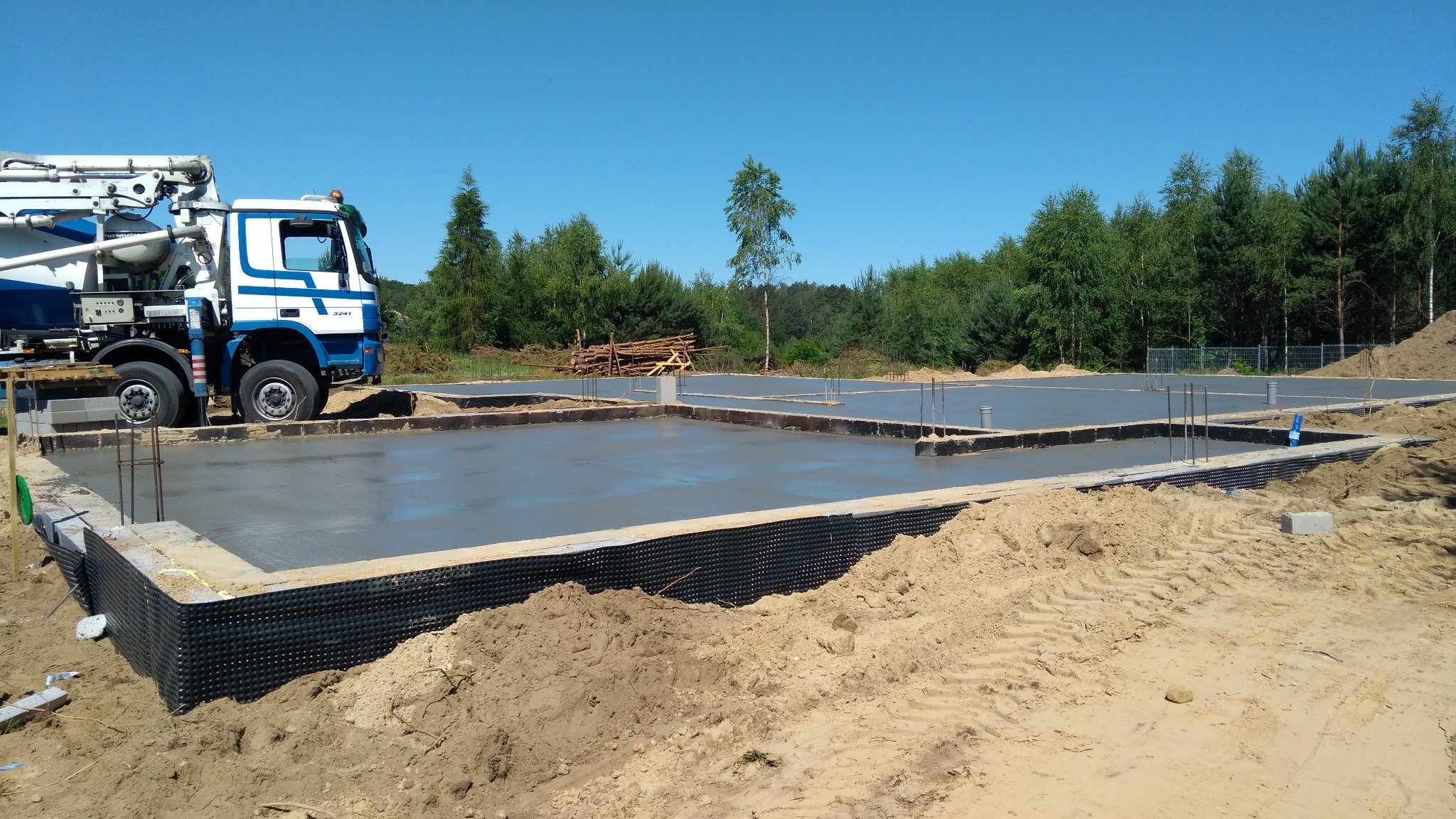Wylany fundament pod budowę domu w Bolszewie, widoczny betonowy podkład, zbrojenie i hydroizolacja, w tle betoniarka i zalesiony teren.