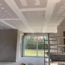 Montaż płyt kartonowo-gipsowych na suficie w pomieszczeniu z dużym oknem, widoczne przewody elektryczne i rusztowanie.