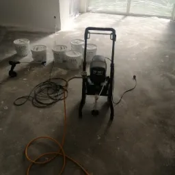 Przygotowania do malowania: agregat malarski PP 90 z pomarańczowym wężem, mieszadło elektryczne, sześć wiader farby oraz zabezpieczone folią okno w pomieszczeniu w trakcie remontu.