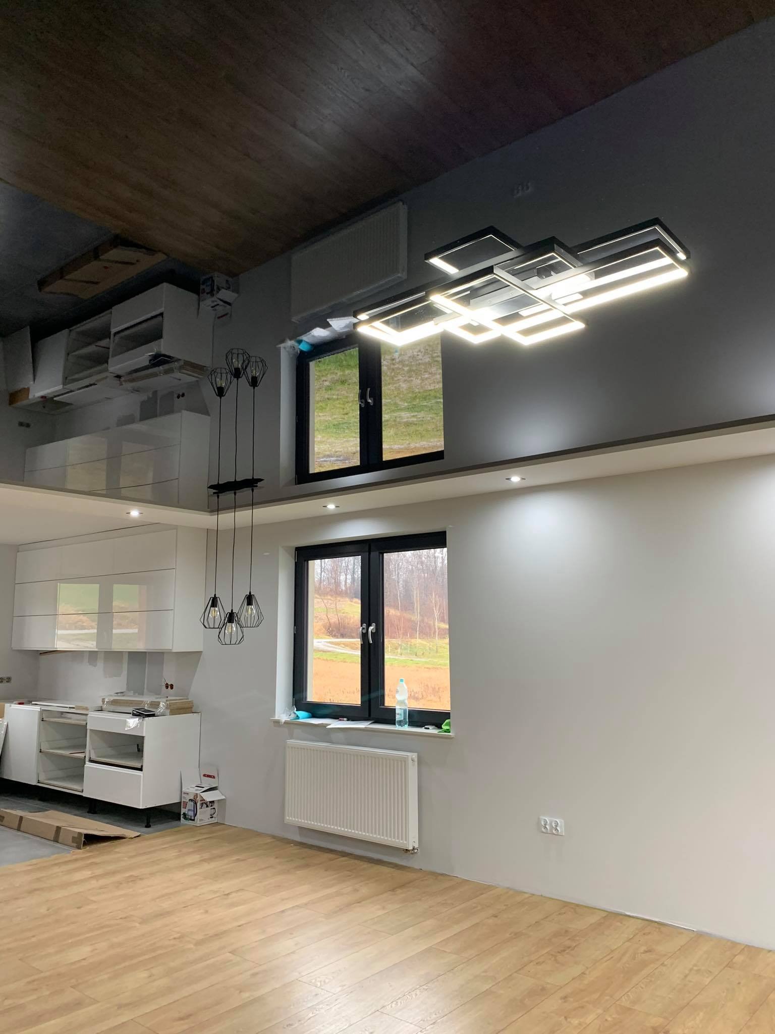 Wnętrze po remoncie: jasne panele podłogowe, białe meble kuchenne, grzejnik, dwa okna z czarnymi ramami, nowoczesna lampa wisząca i sufit z połyskiem odbijający obraz kuchni.