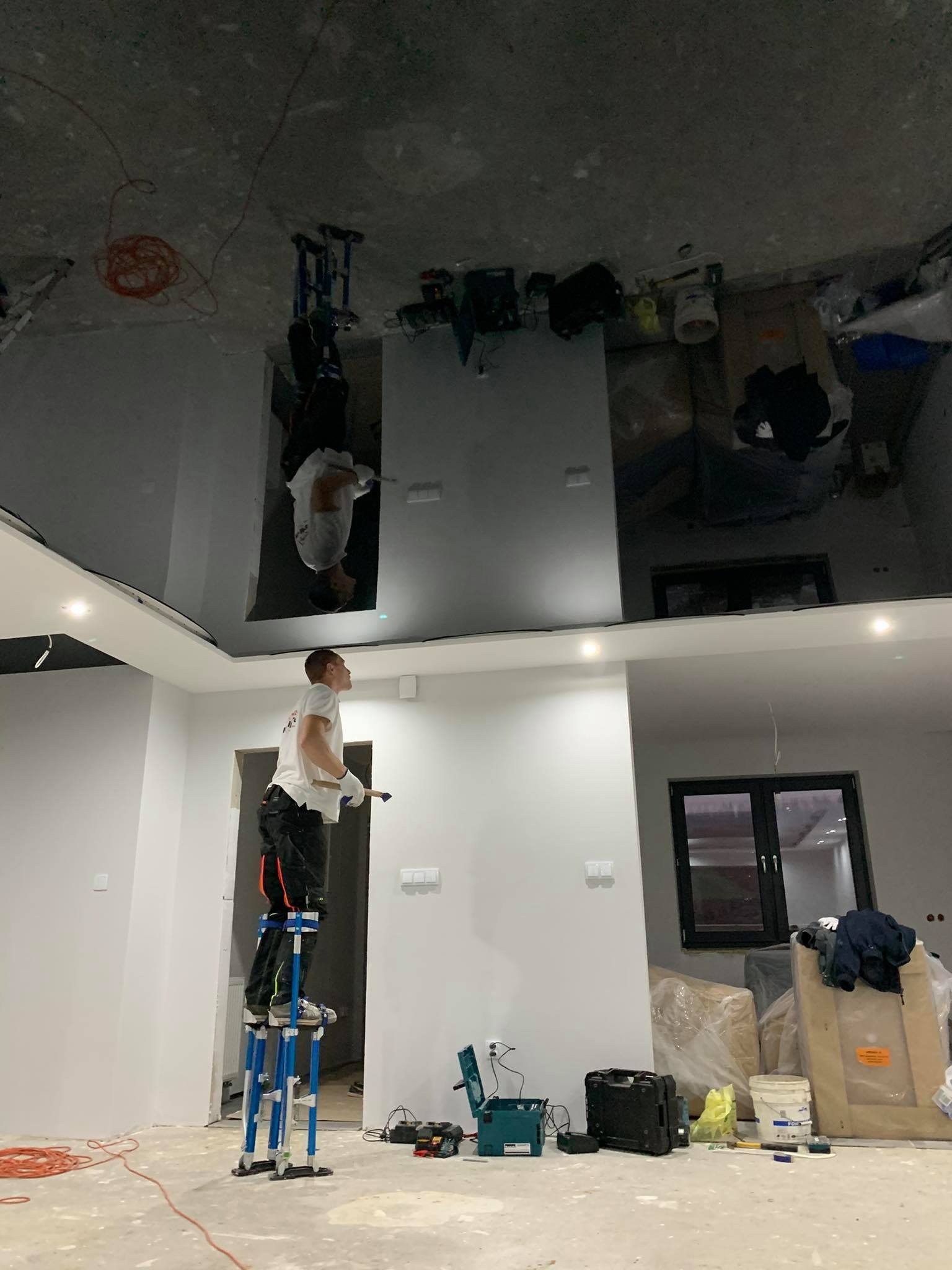 Pracownik na szczudłach maluje sufit w pomieszczeniu z zamontowanym błyszczącym sufitem napinanym. W tle widoczne narzędzia i materiały budowlane.