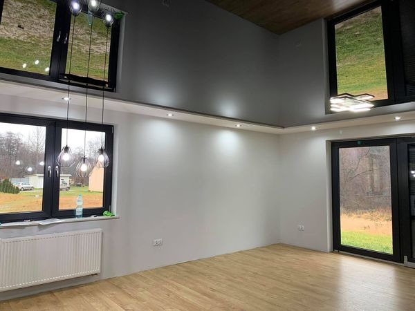 Jasne wnętrze po remoncie: świeżo pomalowane ściany w odcieniach szarości i bieli, drewnopodobna podłoga, czarne ramy okien i drzwi balkonowych, nowoczesne lampy wiszące i sufitowe, grzejnik...