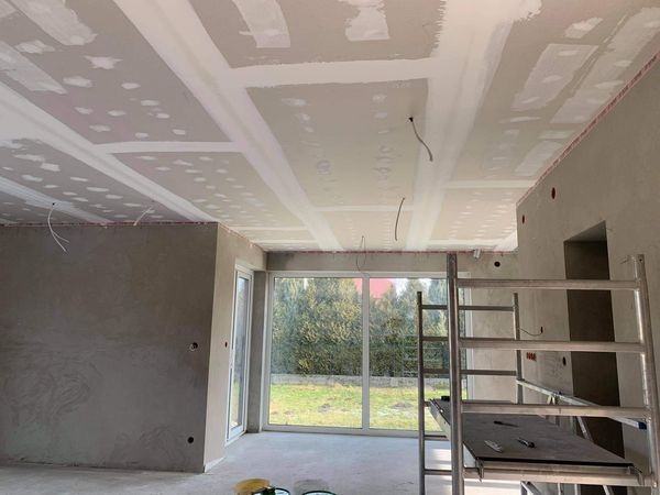 Montaż płyt kartonowo-gipsowych na suficie w pomieszczeniu z dużym oknem, widoczne przewody elektryczne i rusztowanie.