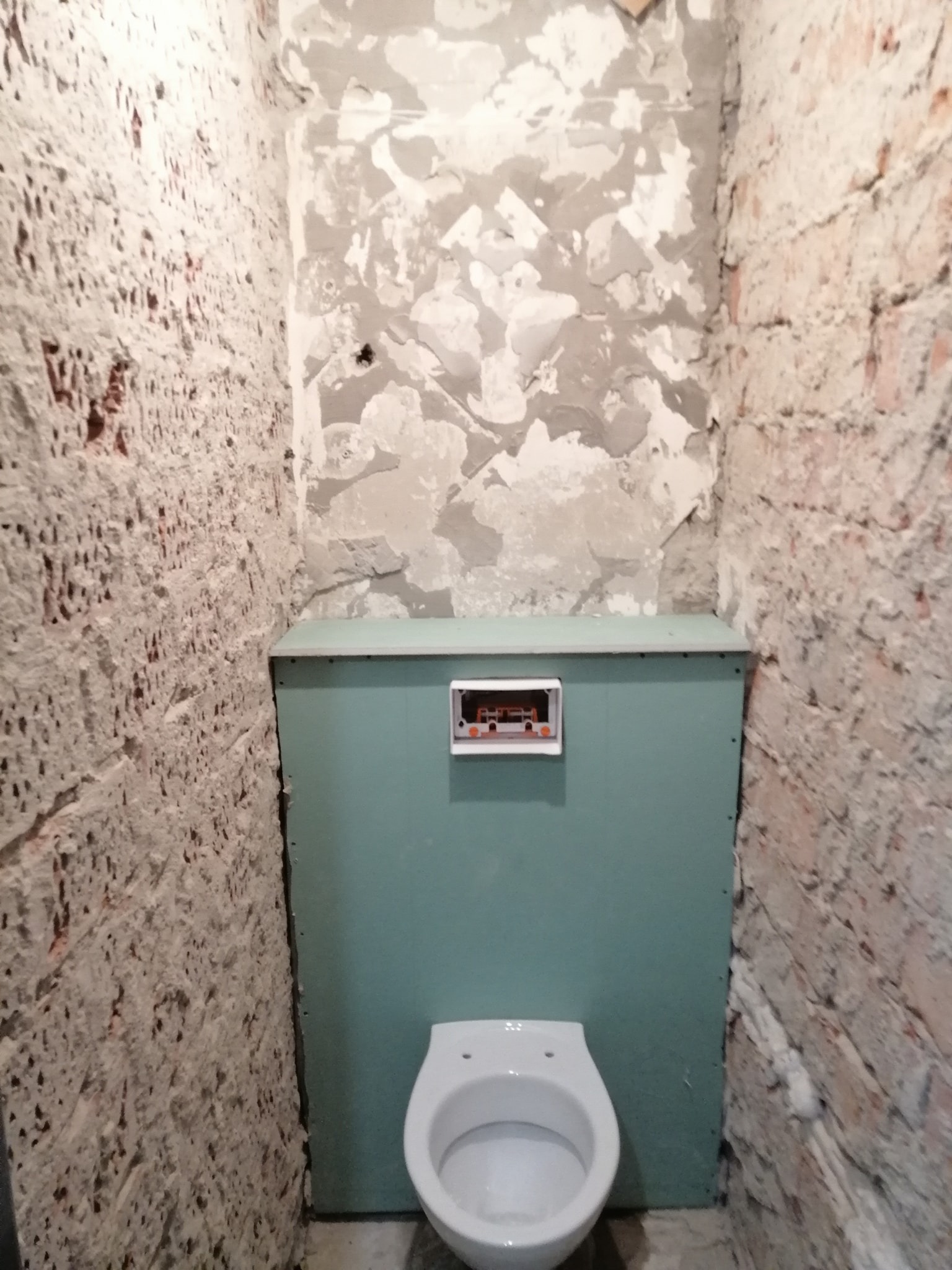 Wnętrze łazienki w trakcie remontu: widoczny stelaż podtynkowy z płytą w kolorze morskim, surowe ściany z fragmentami starej cegły i resztkami zaprawy, oraz biała miska WC.