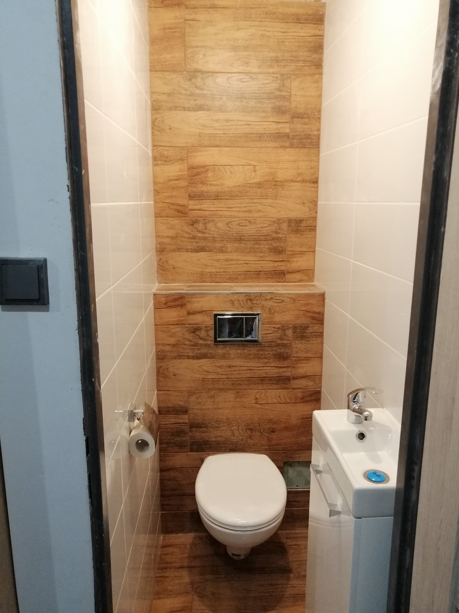 Mała toaleta w wąskiej wnęce, wykończona płytkami imitującymi drewno oraz białymi płytkami, z wiszącą muszlą klozetową i wąską umywalką z szafką, uchwyt na papier toaletowy po lewej stronie.