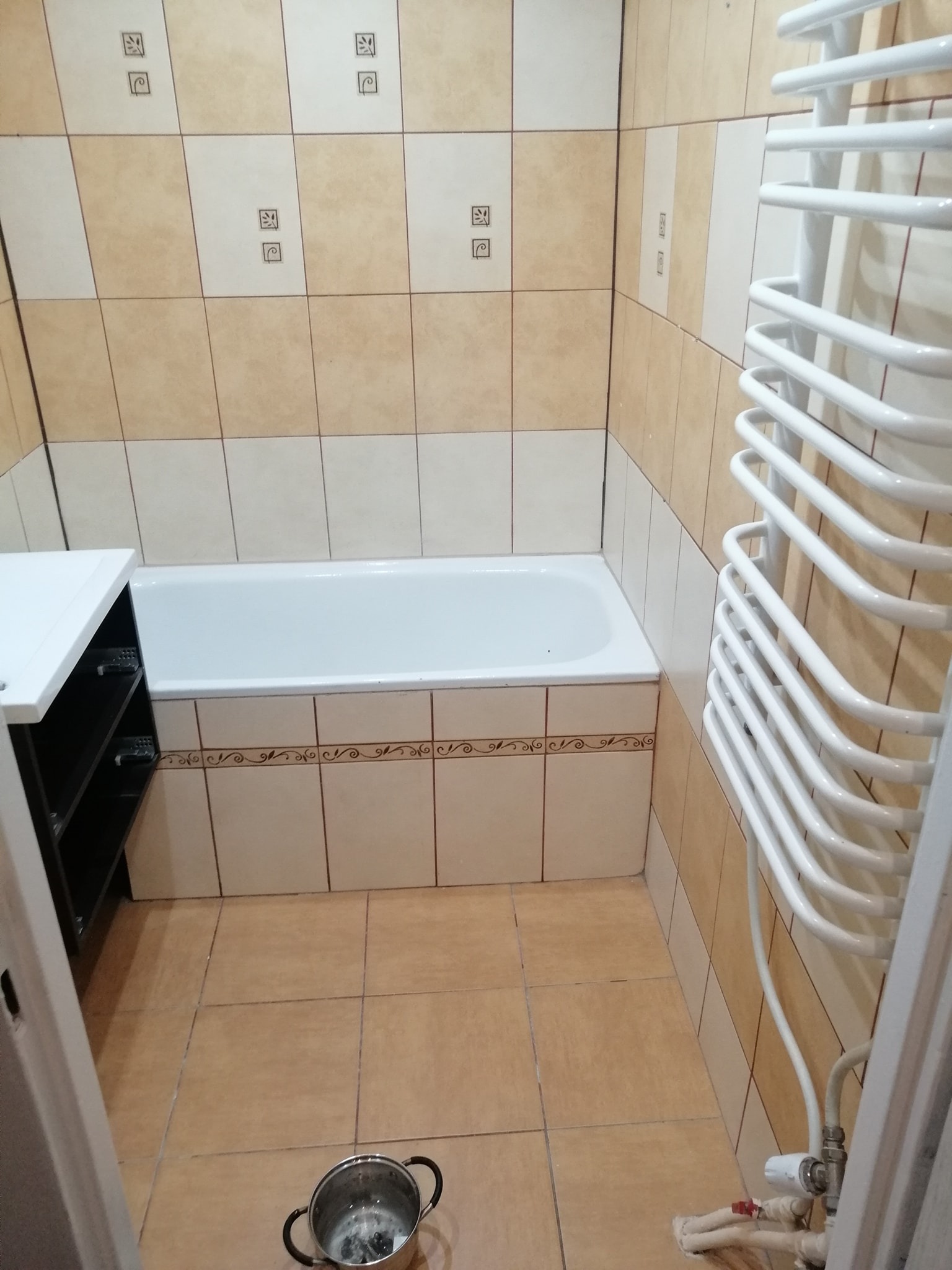 Wykończona łazienka w Woli Żyrakowskiej z beżowo-białymi płytkami, białą wanną i grzejnikiem drabinkowym; na podłodze stoi metalowy garnek.