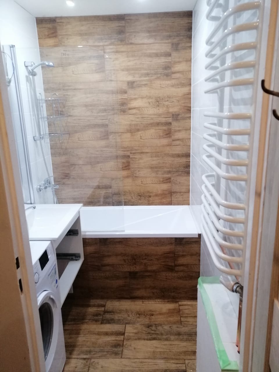 Nowoczesna łazienka z wanną obudowaną płytkami imitującymi drewno, białą umywalką z szafką, pralką i grzejnikiem drabinkowym.