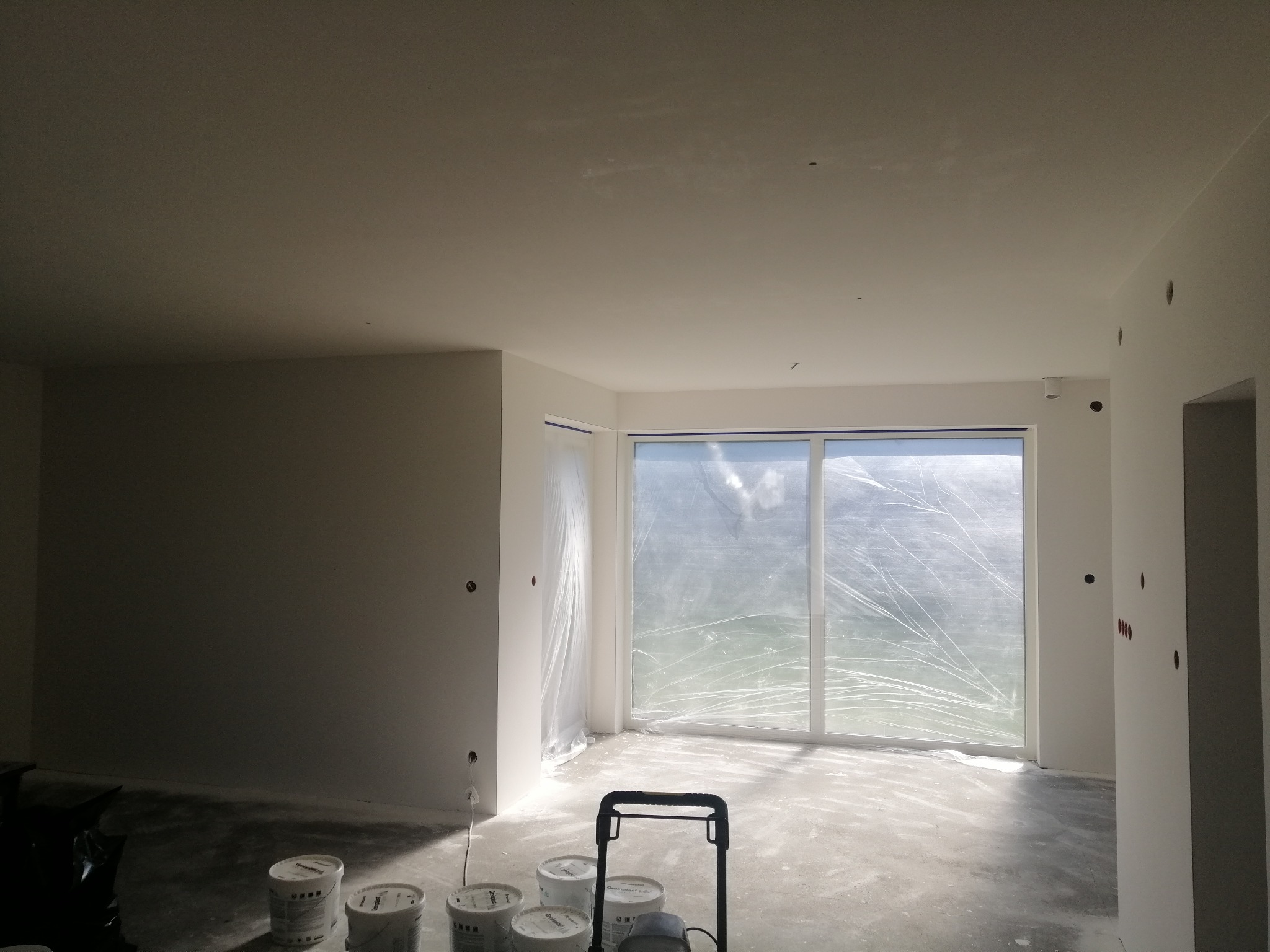 Wnętrze w trakcie przygotowań do malowania: ściany i sufit po szpachlowaniu, okna zabezpieczone folią, widoczne puszki z farbą i wózek malarski na podłodze.