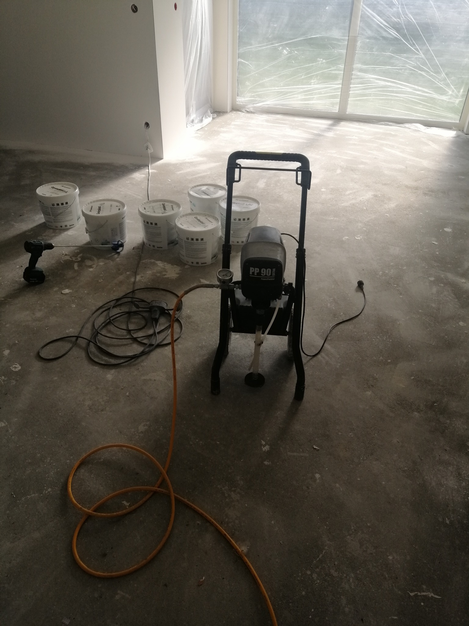 Przygotowania do malowania: agregat malarski PP 90 z pomarańczowym wężem, mieszadło elektryczne, sześć wiader farby oraz zabezpieczone folią okno w pomieszczeniu w trakcie remontu.