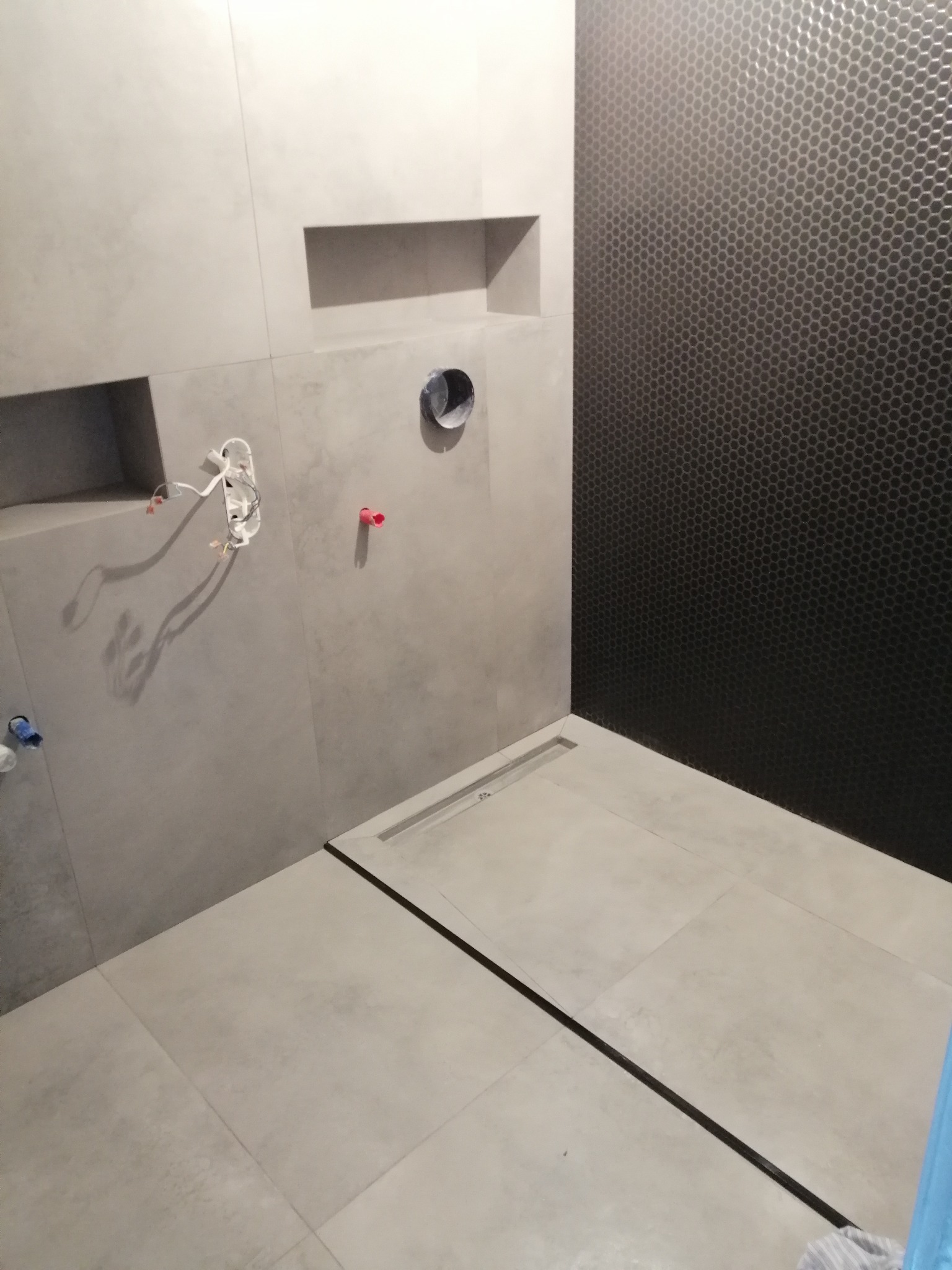 Wnętrze łazienki w trakcie prac wykończeniowych: szare płytki wielkoformatowe na ścianach z wnękami, czarna ściana wyłożona mozaiką w kształcie plastra miodu, widoczne wystające przewody...