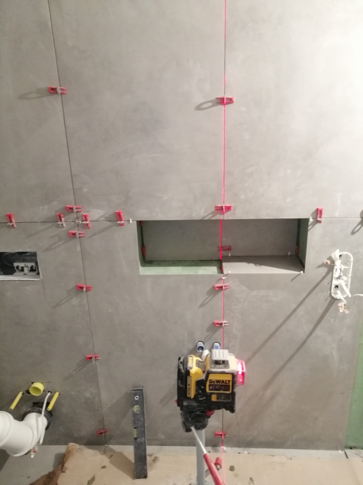 Świeżo ułożone, szare płytki na ścianie łazienki, wyrównywane za pomocą poziomicy laserowej DeWalt i czerwonych klipsów dystansowych, widoczny otwór w ścianie oraz fragment instalacji kanalizacyjnej.