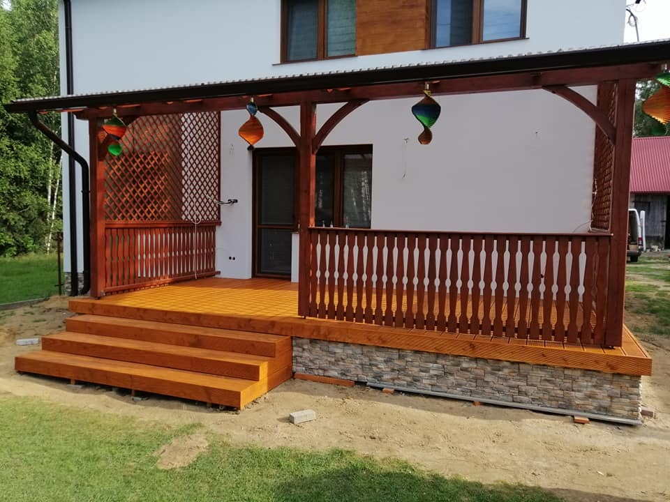 Zadaszony drewniany taras z ażurową balustradą i schodami, wykończony kamienną okładziną, z kolorowymi lampionami wiszącymi pod sufitem, na tle białej ściany domu.