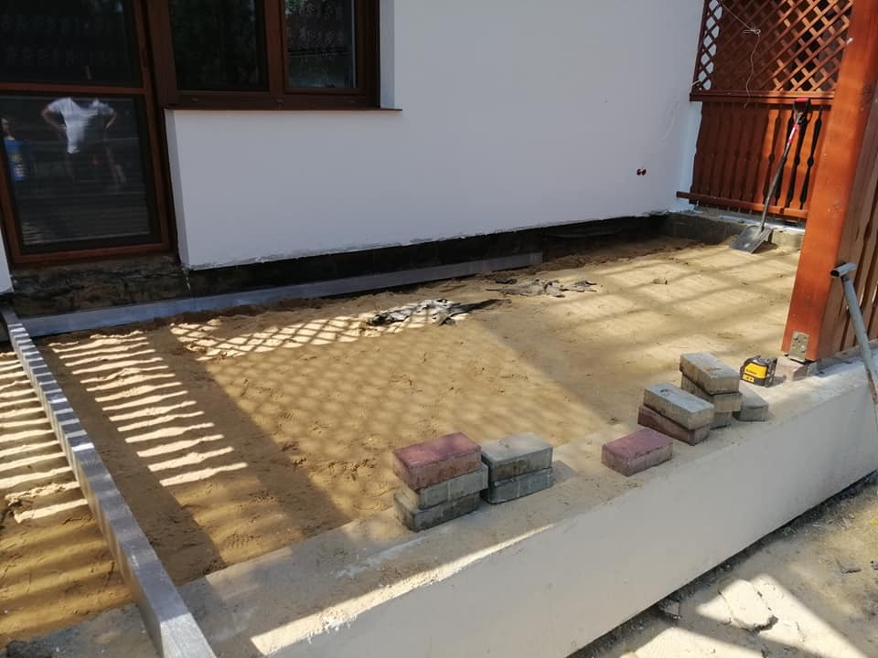 Przygotowanie podłoża pod taras z kostki brukowej, widoczny piasek, obrzeża betonowe i ułożone kostki w kolorach szarości i czerwieni, w tle ściana budynku z oknem.
