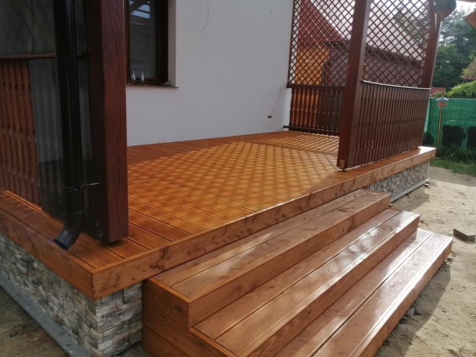 Drewniany taras z ażurową konstrukcją, widoczne schody i podmurówka z kamienia dekoracyjnego, brązowe elementy drewniane kontrastują z białą ścianą budynku.