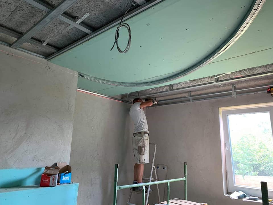 Pracownik na drabinie montuje podwieszany sufit z płyty gipsowo-kartonowej o nietypowym, zaokrąglonym kształcie, widoczna konstrukcja metalowa i laserowy poziom na ścianie, w tle okno i częściowo...