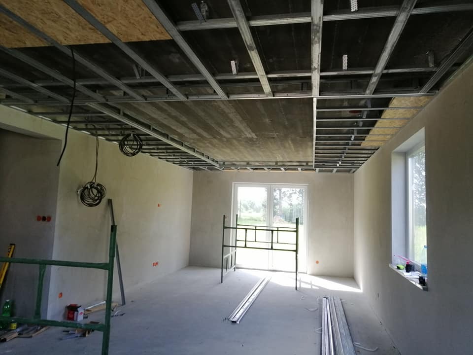 Montaż metalowej konstrukcji sufitu podwieszanego z częściowo zamontowaną płytą OSB w surowym wnętrzu z oknami i rusztowaniem.