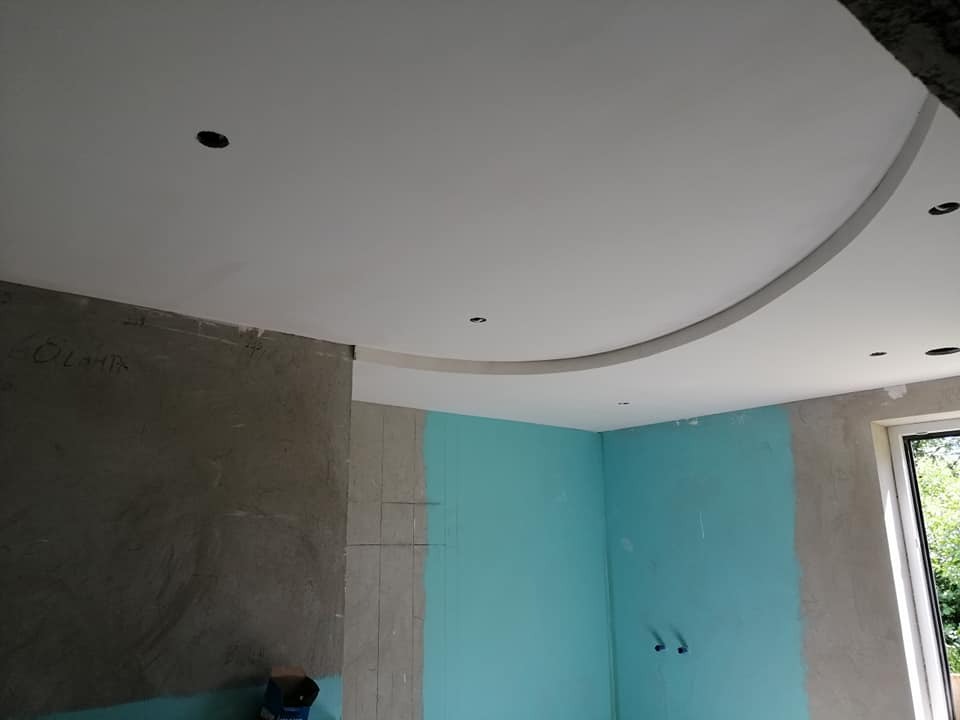 Wnętrze w trakcie prac wykończeniowych: sufit podwieszany z łukowym elementem i punktowym oświetleniem, ściany częściowo pomalowane na turkusowo, fragment ściany w stanie surowym z widocznymi...