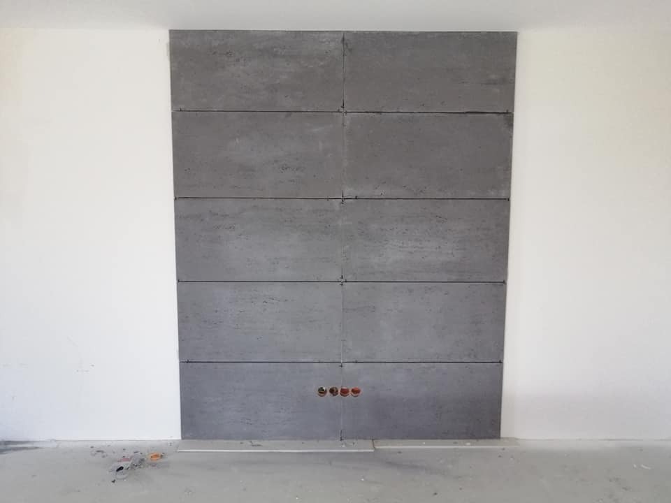 Ściana z szarych paneli imitujących beton, z widocznymi liniami podziału i otworami na gniazdka elektryczne, przylegająca do białej ściany, w trakcie montażu.