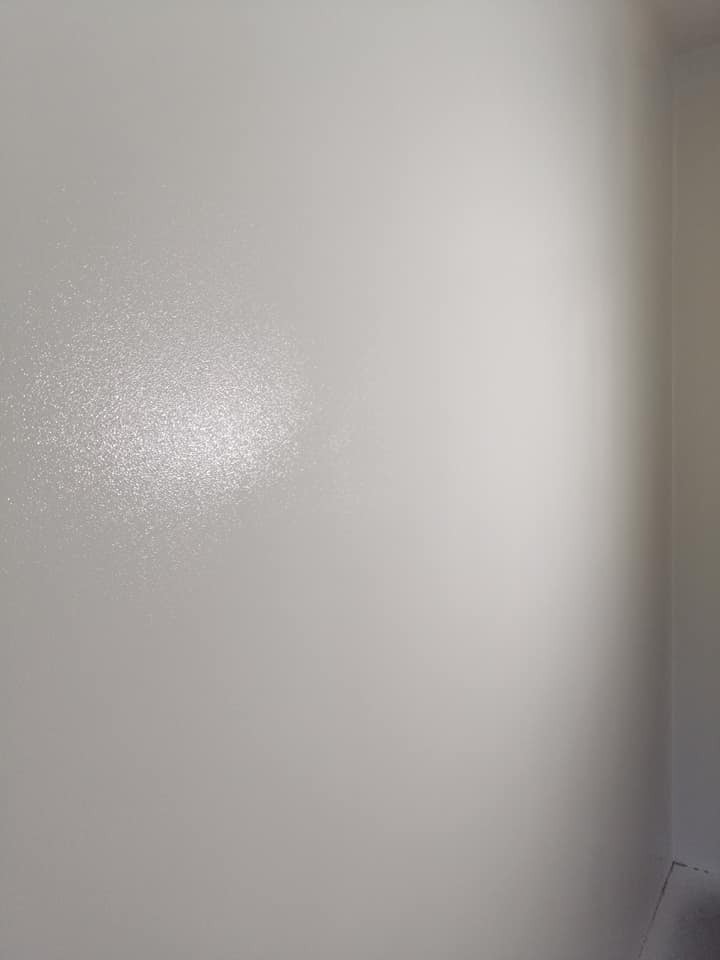 Świeżo pomalowana ściana w odcieniu jasnej szarości z delikatnym połyskiem, widoczny fragment narożnika pomieszczenia, oświetlenie podkreśla strukturę farby.