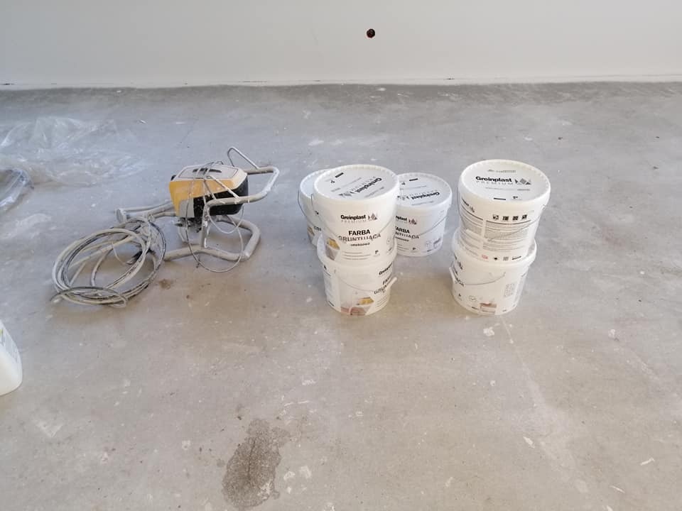 Agregat malarski leżący obok wiader z farbą gruntującą i farbą na betonowej podłodze w trakcie prac malarskich.
