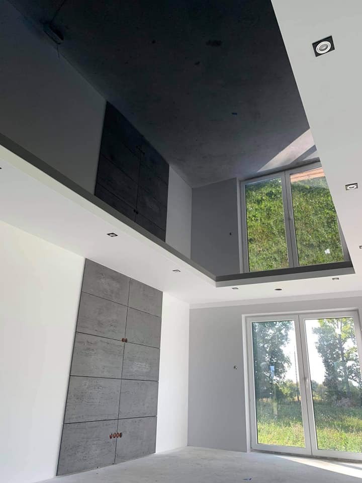 Wnętrze w trakcie wykańczania: Ściana z dekoracyjnych paneli imitujących beton, ciemny sufit z wpuszczonym oświetleniem, duże okna z widokiem na zieleń, widoczne niedoskonałości na podłodze.