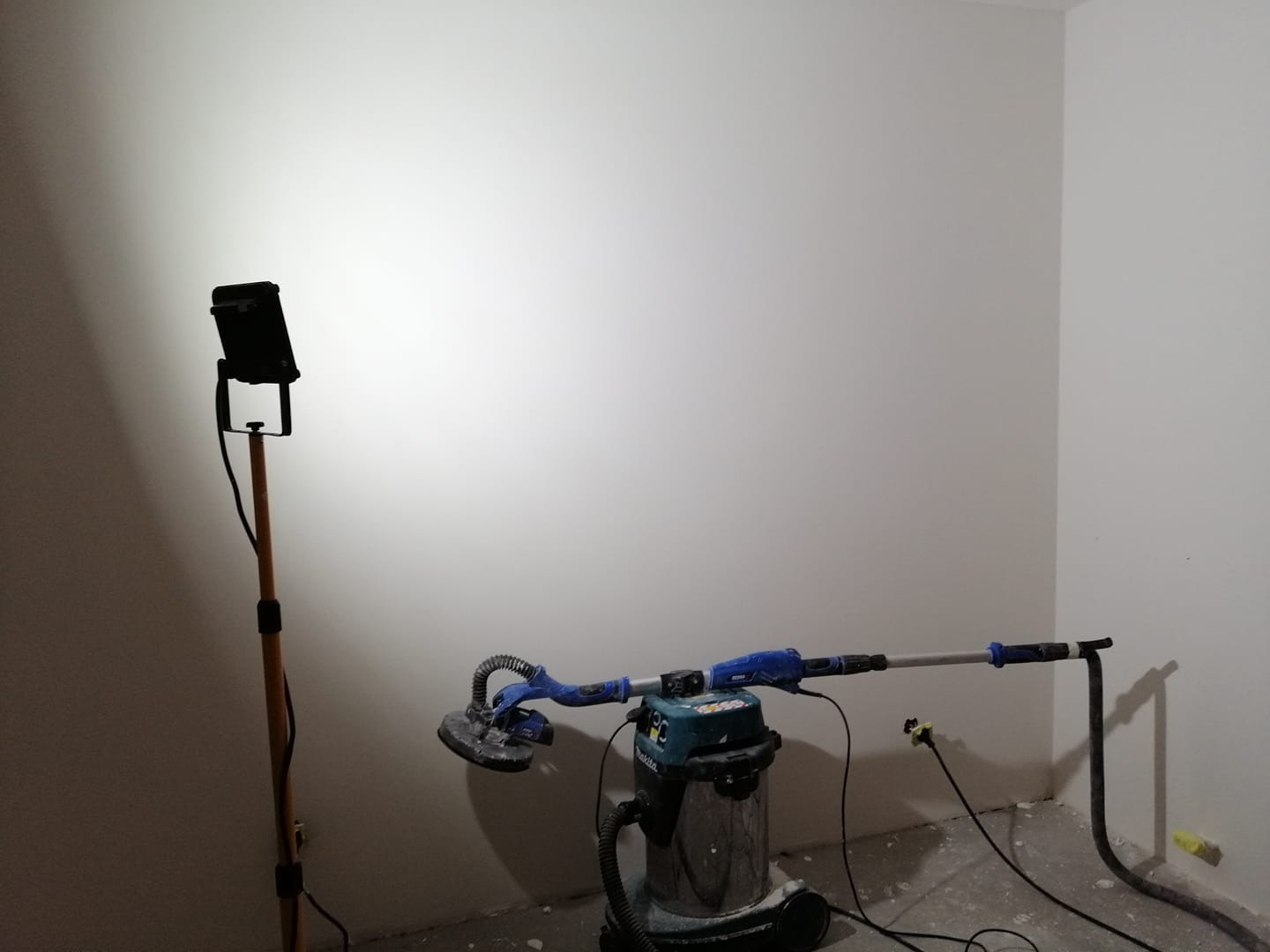 Szlifowanie ściany szlifierką z odkurzaczem przemysłowym i oświetleniem halogenowym na statywie, widoczne ślady pyłu na podłodze.