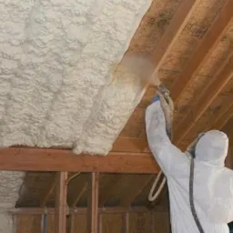 Pracownik w kombinezonie ochronnym aplikuje izolację natryskową na poddaszu, widoczne drewniane elementy konstrukcji dachu.